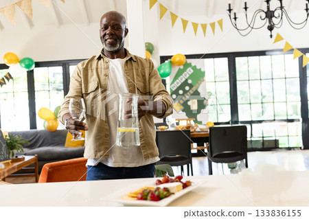 Senior African American man holding pitcher and wine glass by cheese platter, balloons at home Senior African American man holding pitcher and wine glass by cheese platter, balloons at home 133836155