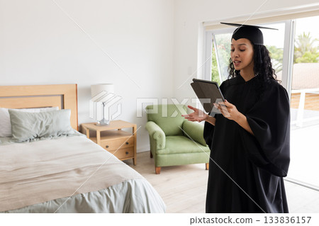 Teenage girl wearing mortarboard and gown reading from tablet in bedroom with armchair, copy space Teenage girl wearing mortarboard and gown reading from tablet in bedroom with armchair, copy space 133836157