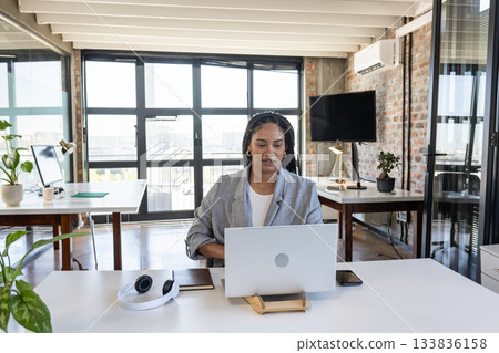African American woman wearing blazer using laptop on stand at desk in open office with headphones 133836158