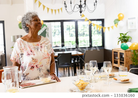 Senior African American woman standing at island in dining area with wine glasses banners balloons 133836160