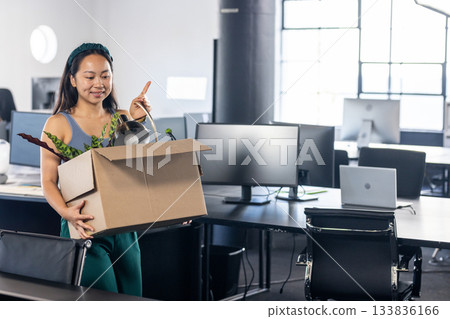 Chinese woman walking through office with moving box containing potted plant, desk lamp, copy space Chinese woman walking through office with moving box containing potted plant, desk lamp, copy space 133836166