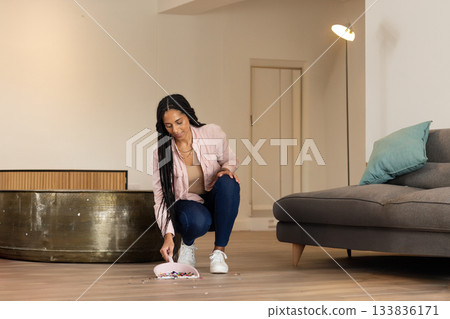 African American woman crouching in living room gathering beads with pink dustpan and brush African American woman crouching in living room gathering beads with pink dustpan and brush 133836171