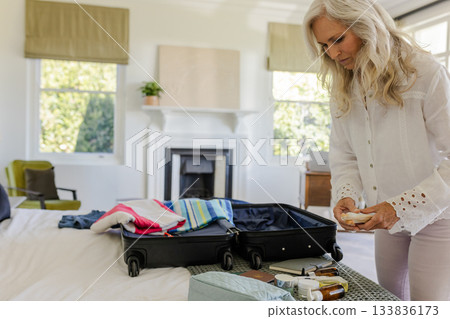 Senior woman packing black suitcase with towels and toiletry bottles in sunlit bedroom, copy space 133836173
