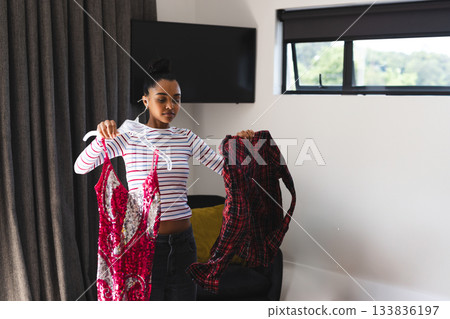 Woman comparing sequined dress and plaid shirt near window in living room with hangers Woman comparing sequined dress and plaid shirt near window in living room with hangers 133836197