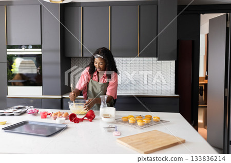 African American woman in green apron mixing batter in glass bowl at kitchen island with muffins 133836214