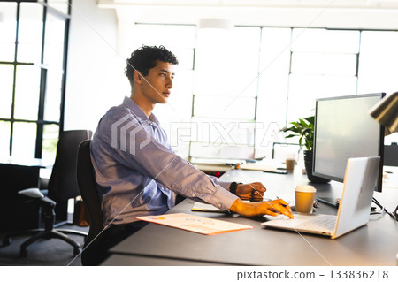 Man in his 30s typing on laptop in modern office with monitor, coffee cup, copy space 133836218