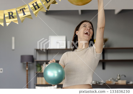 Woman in her 20s holding teal balloon touching gold balloon under birthday banner in living area Woman in her 20s holding teal balloon touching gold balloon under birthday banner in living area 133836230