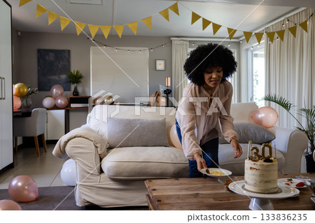 Mid adult African American woman bending toward table at home placing snacks beside birthday cake 133836235