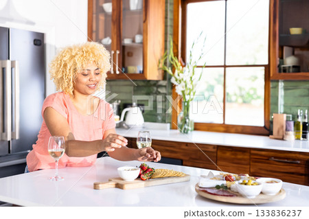 Woman in pink top arranging charcuterie on cutting board beside wine glasses in modern kitchen Woman in pink top arranging charcuterie on cutting board beside wine glasses in modern kitchen 133836237