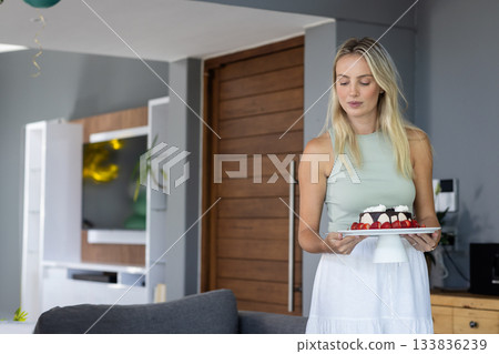 Woman in mid twenties holding strawberry cake inside living room with green balloon, copy space 133836239