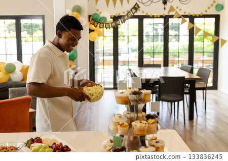 African American man holding popcorn bowl at home counter near cupcakes with balloons, copy space 133836245