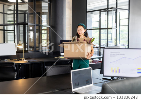 Asian woman holding cardboard box containing potted plant and files while walking through office 133836248