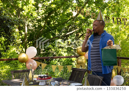 African American man standing on wooden deck holding green gift bag blowing party horn, copy space African American man standing on wooden deck holding green gift bag blowing party horn, copy space 133836250