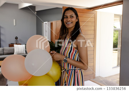 Smiling woman holding peach white balloon cluster by open wooden door at home wearing striped dress 133836256
