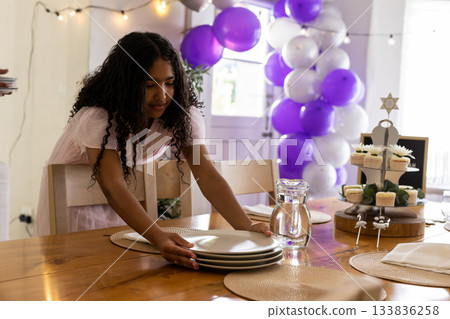 Girl leaning at dining table placing stack of plates among cupcakes and balloon arch, copy space 133836258