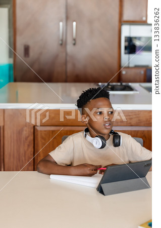 African American school-aged boy studying on tablet with notebook and headphones at kitchen table African American school-aged boy studying on tablet with notebook and headphones at kitchen table 133836262