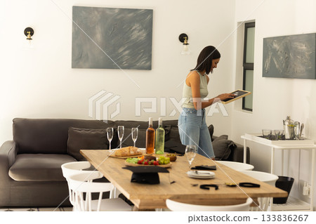 Woman standing by dining table in home with wine, fruit, inspecting picture frame, copy space Woman standing by dining table in home with wine, fruit, inspecting picture frame, copy space 133836267