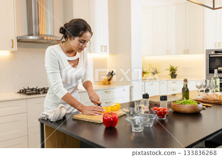 Woman slicing yellow pepper on cutting board at black countertop island with glass bowls, bread Woman slicing yellow pepper on cutting board at black countertop island with glass bowls, bread 133836268