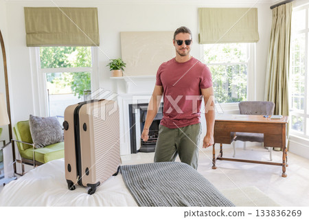 Male traveler standing in guest bedroom beside rolling suitcase and bed with writing desk Male traveler standing in guest bedroom beside rolling suitcase and bed with writing desk 133836269