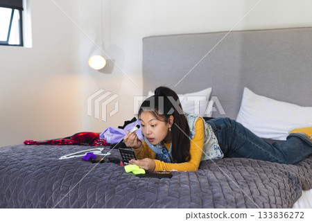 Asian woman applying makeup while lying on quilted bed surrounded by palette, brushes and clothing 133836272