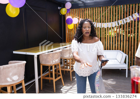 African American woman standing under pergola holding smartphone, party ribbon and birthday banner 133836274