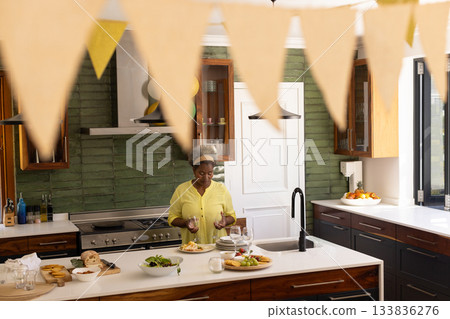 Senior African American woman plating appetizers and arranging wine glasses on island in kitchen 133836276