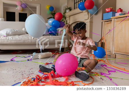 African American male child sitting among balloons, streamers, and confetti on living room floor 133836289