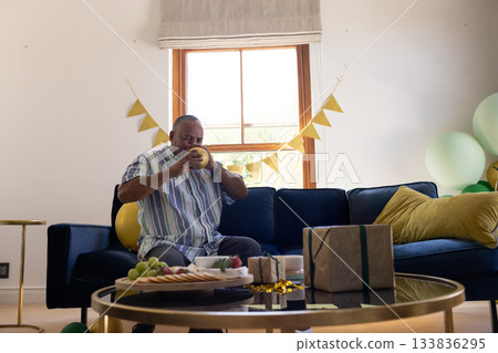 Senior man sitting on dark blue sofa in living room inflating yellow balloon beside gift box 133836295