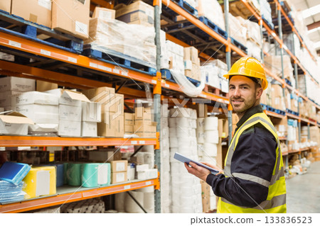 Male worker wearing hard hat and hi-vis vest inspecting stock with tablet in warehouse, copy space 133836523
