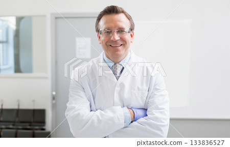 Male doctor standing arms crossed in clinical room wearing lab coat safety glasses and blue gloves Male doctor standing arms crossed in clinical room wearing lab coat safety glasses and blue gloves 133836527