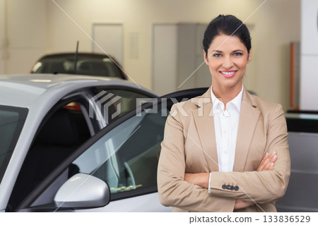 Female professional in blazer standing beside silver sedan with open door in showroom, copy space Female professional in blazer standing beside silver sedan with open door in showroom, copy space 133836529