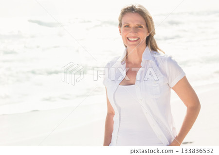 Senior woman smiling while standing on sandy shore in white shirt under bright sunlight, copy space Senior woman smiling while standing on sandy shore in white shirt under bright sunlight, copy space 133836532
