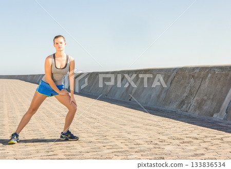 Woman doing side lunge in sportswear, wearing running shoes by sea wall on walkway, copy space 133836534