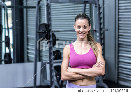 Mid adult woman smiling and standing with arms crossed in gym training area near squat rack 133836536