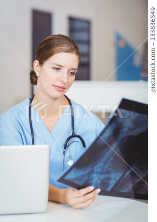 Female doctor wearing scrubs and stethoscope, holding X-ray film, checking laptop at clinic desk Female doctor wearing scrubs and stethoscope, holding X-ray film, checking laptop at clinic desk 133836549