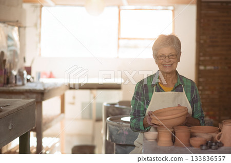Senior woman wearing apron holding clay bowl at pottery wheel smiling in studio, copy space Senior woman wearing apron holding clay bowl at pottery wheel smiling in studio, copy space 133836557