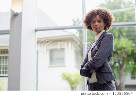 African American woman standing with arms crossed in atrium by glass windows, in suit, copy space 133836564