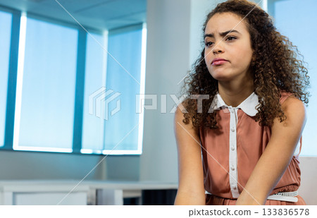 Woman in rust-colored dress sitting leaning forward on white desk in office with blinds, copy space 133836578