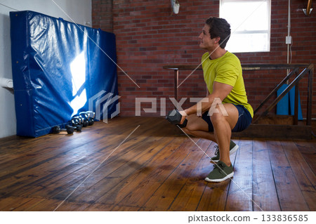 Man wearing green shirt performing weighted squat holding dumbbell in gym with blue mat, copy space 133836585