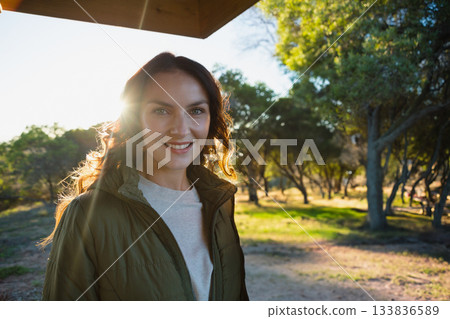 Woman smiling and standing beneath wooden canopy in wooded park wearing green quilted jacket 133836589