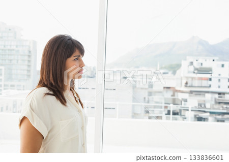 Woman wearing necklace, gazing out large window inside apartment at skyline, copy space Woman wearing necklace, gazing out large window inside apartment at skyline, copy space 133836601