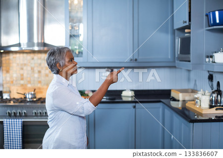 Senior Asian woman pointing at upper cabinet in modern kitchen with stainless steel kettle 133836607