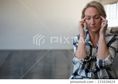 Mature woman sitting pressing hands against temples on bench in room with window, copy space 133836624