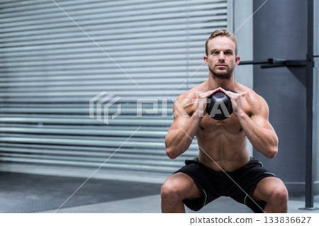 Shirtless man performing kettlebell squat at gym near metal roller shutter door on rubber flooring 133836627
