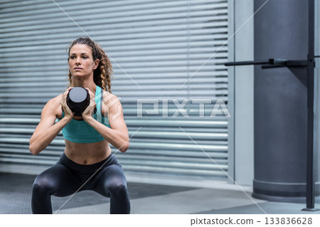 Woman performing weighted squat in gym weight area holding black kettlebell at chest 133836628