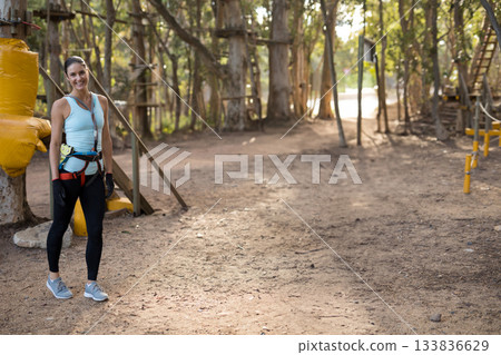 Woman standing at forest obstacle course wearing harness, smiling by rope ladder, copy space Woman standing at forest obstacle course wearing harness, smiling by rope ladder, copy space 133836629