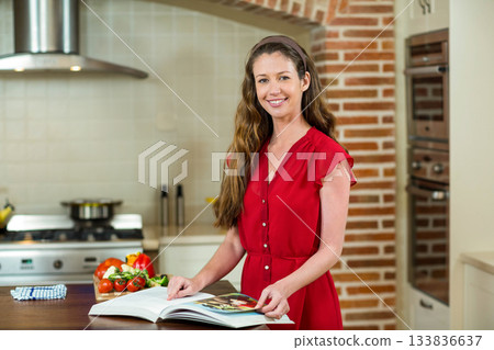 Female home cook wearing red dress reading cookbook on wooden island with tomatoes and bell peppers 133836637