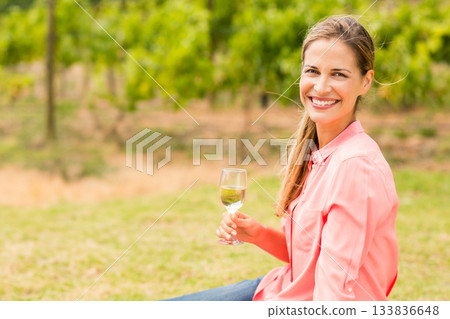 Woman wearing coral-pink shirt, sitting at vineyard edge holding glass of white wine, copy space 133836648