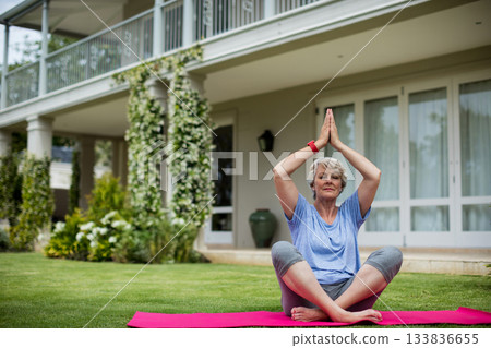 Senior woman sitting on pink yoga mat in front yard wearing smartwatch near potted green vase Senior woman sitting on pink yoga mat in front yard wearing smartwatch near potted green vase 133836655