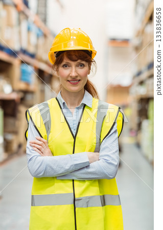 Middle-aged woman standing with arms crossed in warehouse aisle wearing hard hat and safety vest Middle-aged woman standing with arms crossed in warehouse aisle wearing hard hat and safety vest 133836658